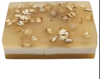 May include: A rectangular bar of handmade soap with a layered design. The top layer is a translucent amber color, embedded with visible oatmeal flakes. The middle layer is white, and the bottom layer is a darker amber color.