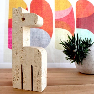 Könnte beinhalten: Eine weiße Steinskulptur eines Pferdes, die auf einem Holztisch steht. Die Skulptur ist abstrakt und hat ein einfaches Design. Das Pferd blickt nach rechts und hat ein Loch im Kopf.