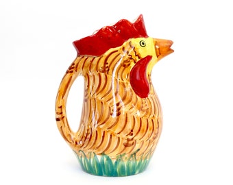Puede incluir: Una jarra de cerámica con forma de gallo con cresta roja, pico amarillo y plumas marrones y naranjas. La jarra tiene un diseño de hierba verde en la base.