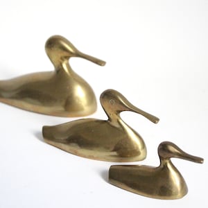 Könnte beinhalten: Drei Messing-Entenfiguren in verschiedenen Größen. Die Enten stehen in einer Reihe, wobei die größte Ente hinten und die kleinste Ente vorne ist. Die Enten sind alle nach rechts im Bild ausgerichtet.