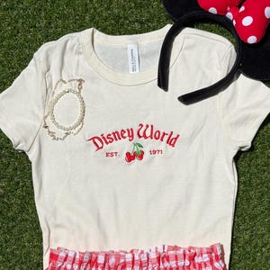 K&ouml;nnte beinhalten: Ein cremefarbenes T-Shirt mit roter "Disney World"-Stickerei und Kirschen. Ein rot-wei&szlig; karierter Rock, ein Minnie-Maus-Ohren-Haarreif und ein goldenes Armband sind ebenfalls im Bild.