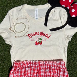 Puede incluir: Camiseta color crema con bordado rojo "Disneyland" y cerezas. Incluye shorts de cuadros rojos y blancos y una diadema con orejas de Minnie. Tambi&eacute;n hay un collar y una pulsera dorados.