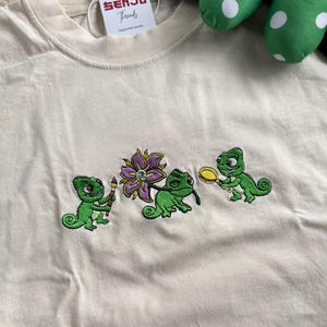 Peut inclure: Un t-shirt couleur crème avec une broderie représentant trois caméléons verts. Un caméléon tient un pinceau, un autre interagit avec une fleur violette et le troisième tient une loupe jaune. Une étiquette blanche indique Senju Threads.