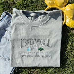 Puede incluir: Una camiseta gris claro con el texto "DISNEY WORLD ESTABLISHED 1971 LAKE BUENA VISTA, FLORIDA" bordado en la parte delantera. El texto est&aacute; en negro con un &aacute;rbol verde, un castillo azul, una bola gris y una cabeza de Mickey Mouse blanca bordada encima.