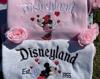 May include: Two sweatshirts, one pink and one gray, embroidered with "Disneyland" in script. The pink sweatshirt features Minnie Mouse, the gray one Mickey Mouse, both with hearts and "Est. 1955." Pink rose accents and a pink bow headband are also visible.