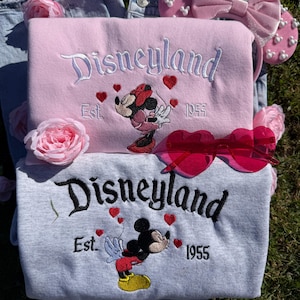 May include: Two sweatshirts, one pink and one gray, embroidered with "Disneyland Est. 1955" and a cartoon character. Pink heart accents and a Minnie Mouse design adorn the pink sweatshirt, while the gray one features Mickey Mouse. Pink heart-shaped sunglasses and a pink bow headband are also present.