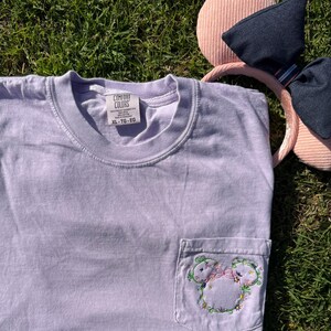 May include: A lavender t-shirt with a pocket featuring an embroidered floral Mickey Mouse design. The t-shirt has a Comfort Colors tag with the size XL - T5 - EG.