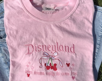 Puede incluir: Camiseta rosa claro de cuello redondo con la palabra "Disneyland" bordada en cursiva. Debajo, un diseño de cerezas con un lazo y la frase "where dreams really do come true". La camiseta está doblada sobre unos vaqueros azules.