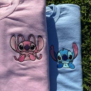 Puede incluir: Dos sudaderas con personajes bordados de Stitch y Angel. La sudadera de la izquierda es rosa con Angel, y la sudadera de la derecha es azul con Stitch.