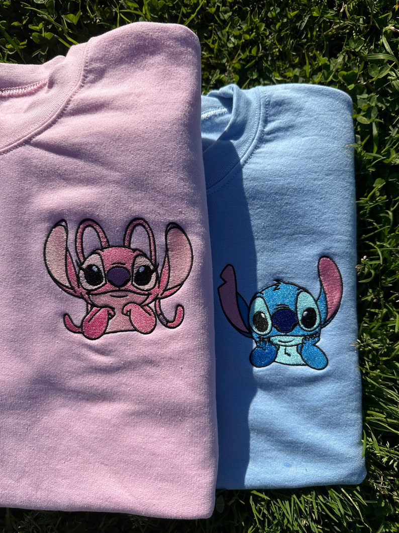 Puede incluir: Dos sudaderas con personajes bordados de Stitch y Angel. La sudadera de la izquierda es rosa con Angel, y la sudadera de la derecha es azul con Stitch.