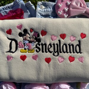 May include: A beige Disneyland sweatshirt with embroidered Mickey and Minnie Mouse, surrounded by red and pink hearts. The word "Disneyland" is embroidered in black. A pink headband with white Mickey Mouse shapes is visible.