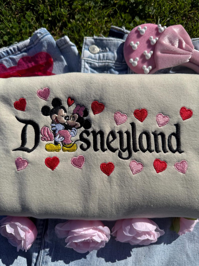 May include: A beige Disneyland sweatshirt with embroidered Mickey and Minnie Mouse, surrounded by red and pink hearts. The word "Disneyland" is embroidered in black. A pink headband with white Mickey Mouse shapes is visible.