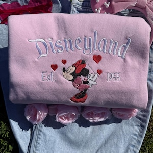 May include: A light pink Disneyland sweatshirt with embroidered text and Minnie Mouse graphic. The design includes red hearts and the year 1955. The sweatshirt is laid on top of blue jeans and surrounded by pink roses.