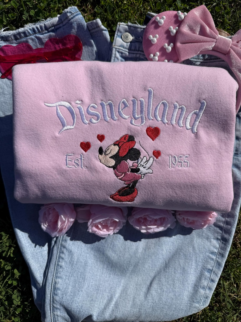 May include: A light pink Disneyland sweatshirt with embroidered text and Minnie Mouse graphic. The design includes red hearts and the year 1955. The sweatshirt is laid on top of blue jeans and surrounded by pink roses.