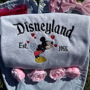 May include: A light gray Disneyland sweatshirt with embroidered black lettering and a Mickey Mouse graphic. The sweatshirt features red heart accents and the text "Est. 1955". Pink roses, a pink bow, and denim jeans are also visible.