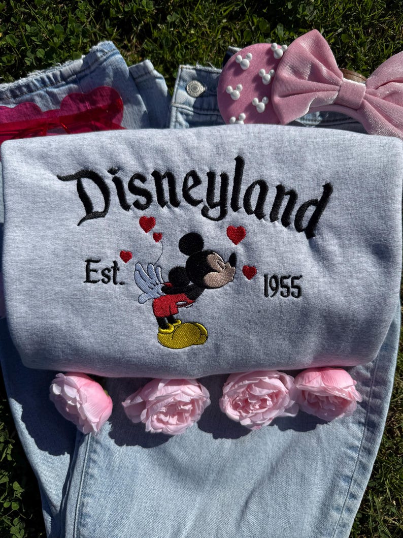 May include: A light gray Disneyland sweatshirt with embroidered black lettering and a Mickey Mouse graphic. The sweatshirt features red heart accents and the text "Est. 1955". Pink roses, a pink bow, and denim jeans are also visible.