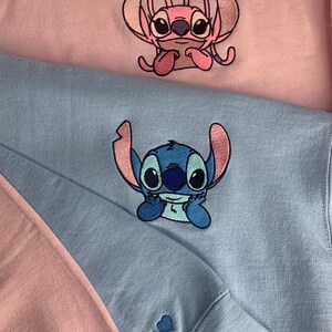 Puede incluir: Dos sudaderas rosas y azules con personajes de Stitch bordados. La sudadera rosa tiene un personaje de Stitch rosa con el texto "J &hearts;" en la manga. La sudadera azul tiene un personaje de Stitch azul con el texto "K &hearts;" en la manga.