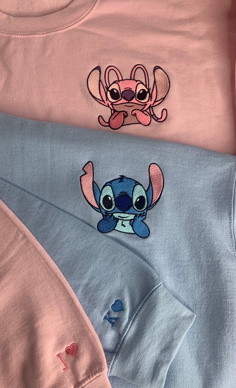 Puede incluir: Dos sudaderas rosas y azules con personajes de Stitch bordados. La sudadera rosa tiene un personaje de Stitch rosa con el texto "J &hearts;" en la manga. La sudadera azul tiene un personaje de Stitch azul con el texto "K &hearts;" en la manga.