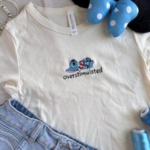 May include: Cream-colored t-shirt with an embroidered blue cartoon character and the word "overstimulated." Light blue denim shorts, a polka-dot bow headband, and blue thread spools are also in the image.