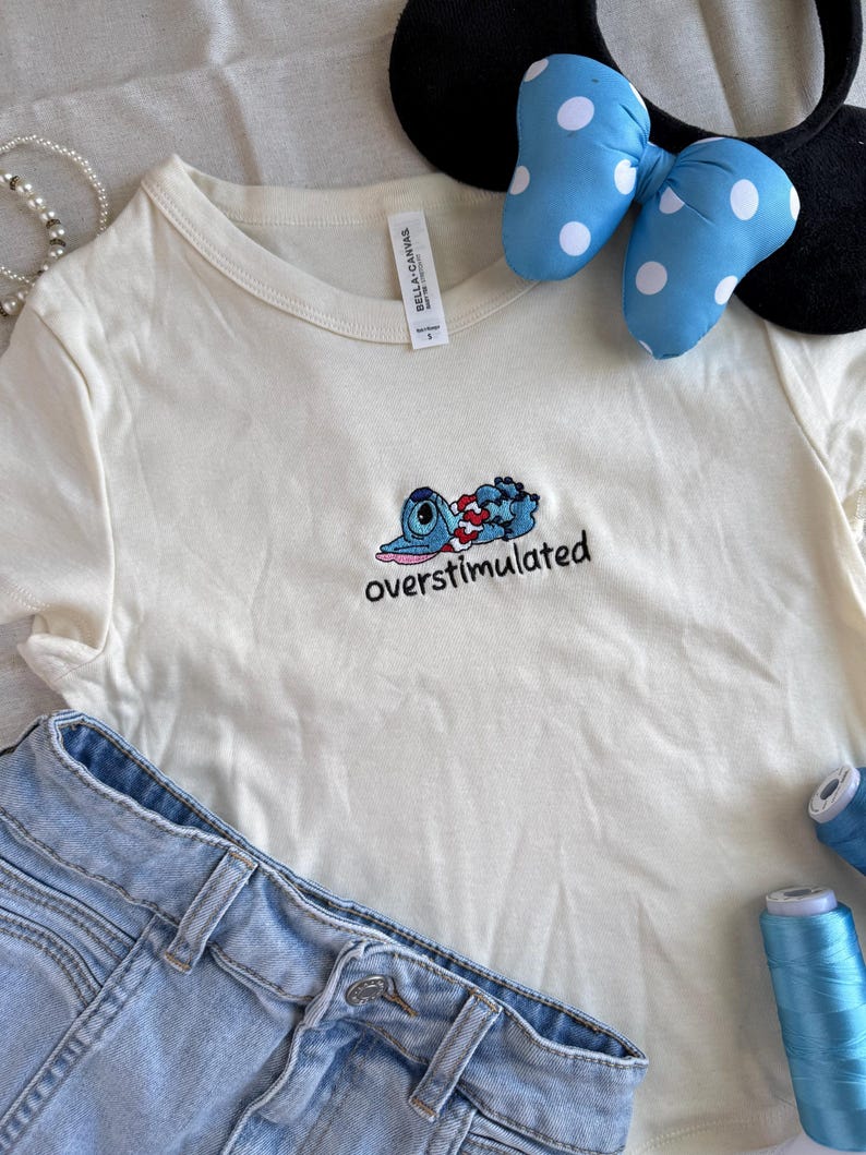 May include: Cream-colored t-shirt with an embroidered blue cartoon character and the word "overstimulated." Light blue denim shorts, a polka-dot bow headband, and blue thread spools are also in the image.