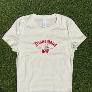 Puede incluir: Camiseta color crema con cuello redondo. La parte delantera presenta la palabra "Disneyland" en escritura roja, con "EST. 1955" debajo, y un par de cerezas rojas. La camiseta est&aacute; hecha de un material suave.