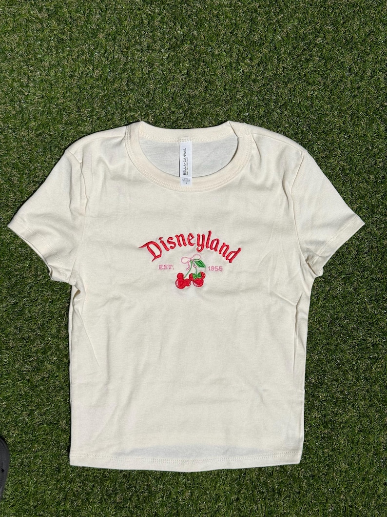 Puede incluir: Camiseta color crema con cuello redondo. La parte delantera presenta la palabra "Disneyland" en escritura roja, con "EST. 1955" debajo, y un par de cerezas rojas. La camiseta est&aacute; hecha de un material suave.