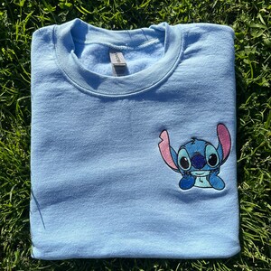Puede incluir: Una sudadera de color azul claro con una peque&ntilde;a imagen bordada de Stitch de Lilo & Stitch. Stitch es azul con orejas rosas y est&aacute; sentado con los brazos cruzados.