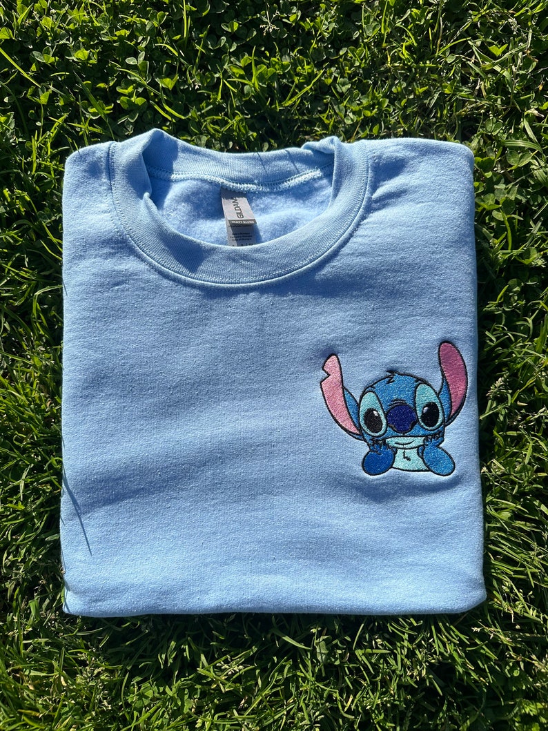 Puede incluir: Una sudadera de color azul claro con una peque&ntilde;a imagen bordada de Stitch de Lilo & Stitch. Stitch es azul con orejas rosas y est&aacute; sentado con los brazos cruzados.