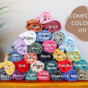 Puede incluir: Una pila de muestras de tela enrolladas en varios colores, incluyendo azul, verde, rojo y amarillo. Las muestras de tela est&aacute;n etiquetadas con nombres como "Berry", "Seafoam", "Watermelon" y "Paprika". El texto "COMFORT COLORS 1717" se muestra encima de las muestras de tela.