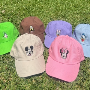 May include: Six baseball caps in different colours, each embroidered with a cartoon character. The caps are green, brown, lavender, light blue, pink, and tan, and are displayed on green grass. The caps are a classic style.
