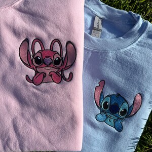 Puede incluir: Dos camisetas con personajes de Stitch bordados. La camiseta rosa tiene un Stitch rosa con un fondo azul claro. La camiseta azul tiene un Stitch azul con un fondo blanco.