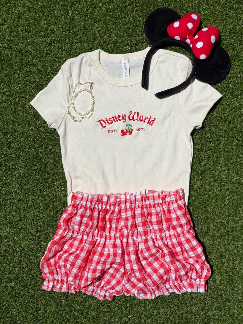 K&ouml;nnte beinhalten: Ein cremefarbenes T-Shirt mit roter "Disney World"-Stickerei und Kirschen. Ein rot-wei&szlig; karierter Rock, ein Minnie-Maus-Ohren-Haarreif und ein goldenes Armband sind ebenfalls im Bild.