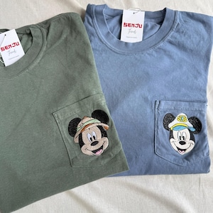 Puede incluir: Una camiseta verde oliva y una azul polvoriento, cada una con un bolsillo en el pecho con una cabeza de Mickey Mouse bordada. La camiseta oliva muestra a Mickey con sombrero de safari, la azul con gorra de capit&aacute;n. Ambas tienen etiquetas Senju Threads.