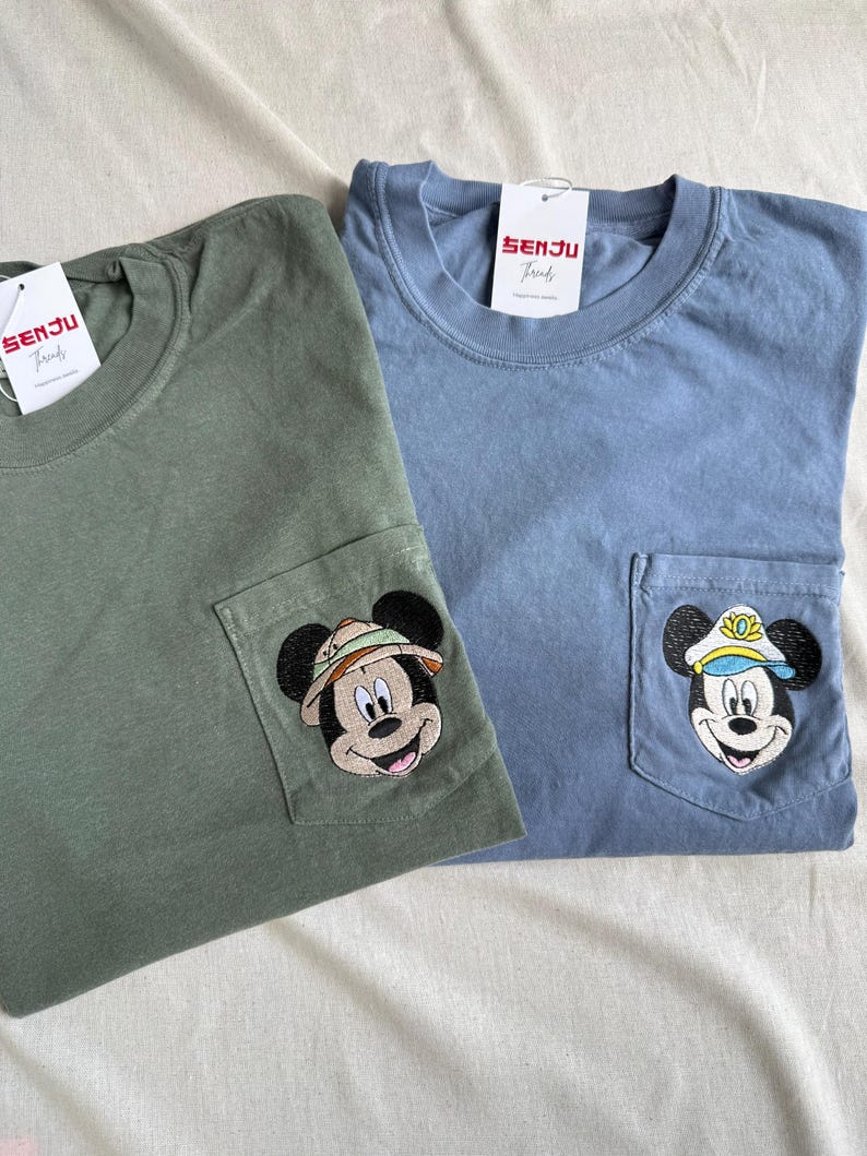 Puede incluir: Una camiseta verde oliva y una azul polvoriento, cada una con un bolsillo en el pecho con una cabeza de Mickey Mouse bordada. La camiseta oliva muestra a Mickey con sombrero de safari, la azul con gorra de capit&aacute;n. Ambas tienen etiquetas Senju Threads.