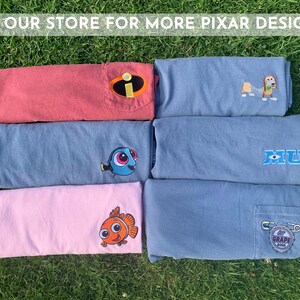 May include: Six folded t-shirts with different Pixar character designs. The shirts are in various colors, including red, blue, and pink. The designs include a character from the movie "Monsters Inc.", a character from the movie "Finding Nemo", and a character from the movie "Toy Story".