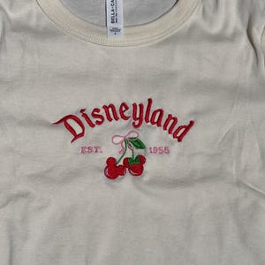 Puede incluir: Camiseta color crema con cuello redondo con bordado rojo "Disneyland" y gr&aacute;fico de cerezas. El texto "EST. 1955" tambi&eacute;n est&aacute; bordado en rosa. La camiseta tiene una etiqueta que dice "BELLA + CANVAS".