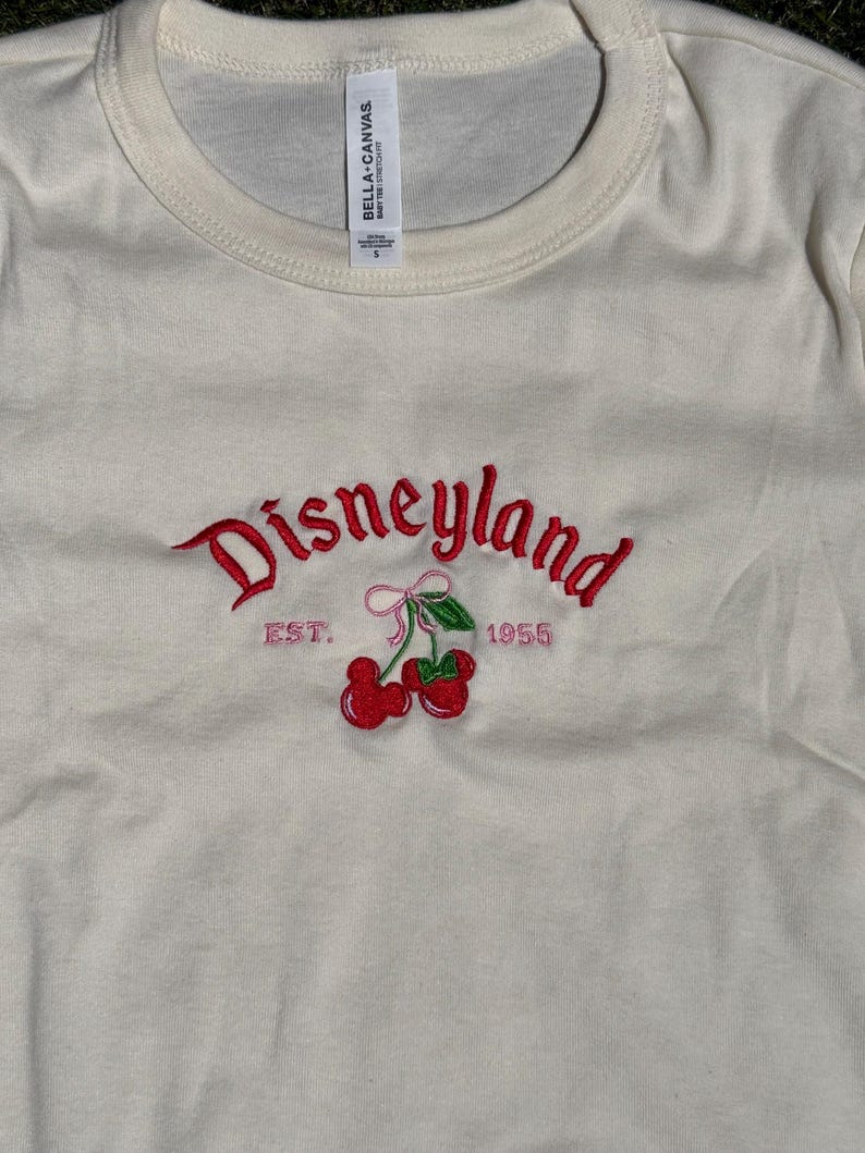 Puede incluir: Camiseta color crema con cuello redondo con bordado rojo "Disneyland" y gr&aacute;fico de cerezas. El texto "EST. 1955" tambi&eacute;n est&aacute; bordado en rosa. La camiseta tiene una etiqueta que dice "BELLA + CANVAS".
