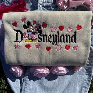 May include: A beige sweatshirt with the word "Disneyland" embroidered in black, featuring Mickey and Minnie Mouse embracing. Red and pink hearts are embroidered around the text. A pink bow headband and pink roses are also visible.