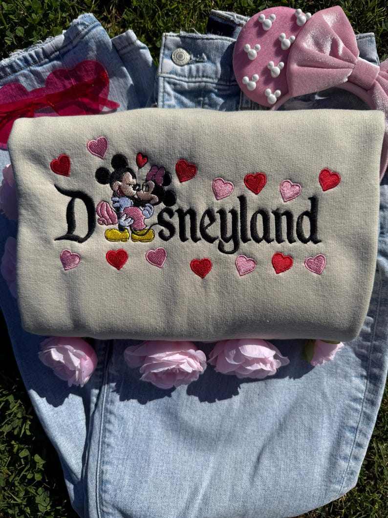 May include: A beige sweatshirt with the word "Disneyland" embroidered in black, featuring Mickey and Minnie Mouse embracing. Red and pink hearts are embroidered around the text. A pink bow headband and pink roses are also visible.