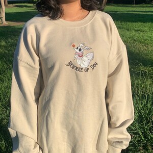 Puede incluir: Una sudadera beige con una imagen bordada de un perro fantasma con una calabaza en la boca. El texto "Beware of Dog" est&aacute; bordado debajo de la imagen.