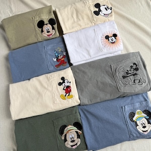Puede incluir: Una colecci&oacute;n de ocho camisetas dobladas en varios colores, incluyendo oliva, crema, blanco, azul y gris. Cada camiseta presenta un bolsillo en el pecho con un dise&ntilde;o bordado de Mickey Mouse, mostrando diferentes atuendos y poses del personaje.