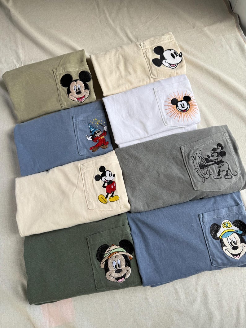 Puede incluir: Una colecci&oacute;n de ocho camisetas dobladas en varios colores, incluyendo oliva, crema, blanco, azul y gris. Cada camiseta presenta un bolsillo en el pecho con un dise&ntilde;o bordado de Mickey Mouse, mostrando diferentes atuendos y poses del personaje.
