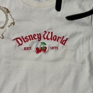 K&ouml;nnte beinhalten: Cremefarbenes T-Shirt mit dem gestickten Schriftzug "Disney World" in roter Schrift, zusammen mit einem Paar Kirschen und dem Jahr "1971". Das Shirt hat einen Rundhalsausschnitt und kurze &Auml;rmel.