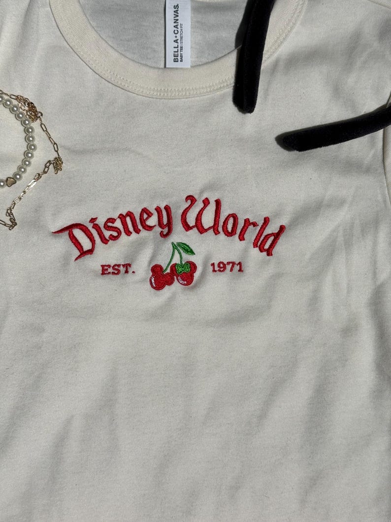 K&ouml;nnte beinhalten: Cremefarbenes T-Shirt mit dem gestickten Schriftzug "Disney World" in roter Schrift, zusammen mit einem Paar Kirschen und dem Jahr "1971". Das Shirt hat einen Rundhalsausschnitt und kurze &Auml;rmel.