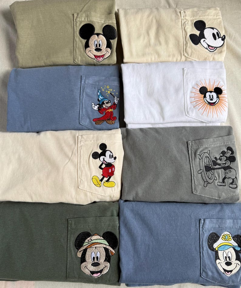 May include: A collection of eight folded t-shirts in various colors including olive, cream, blue, white, and gray. Each shirt features a chest pocket with a unique embroidered Mickey Mouse design, ranging from classic portraits to themed character outfits.