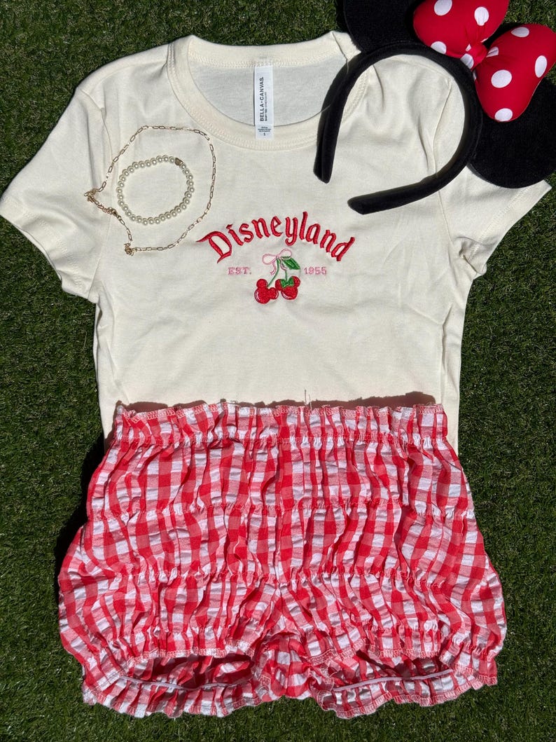Puede incluir: Camiseta color crema con bordado rojo "Disneyland" y cerezas. Incluye shorts de cuadros rojos y blancos y una diadema con orejas de Minnie. Tambi&eacute;n hay un collar y una pulsera dorados.