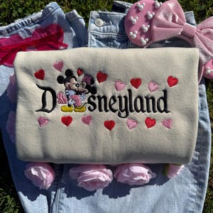May include: A beige Disneyland sweatshirt with embroidered Mickey and Minnie Mouse, surrounded by red and pink hearts. The sweatshirt is laid on top of blue jeans, with pink roses and a pink headband with Mickey Mouse shapes.