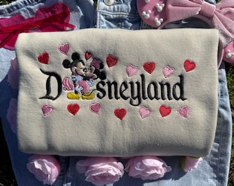 May include: A beige Disneyland sweatshirt with embroidered Mickey and Minnie Mouse, surrounded by red and pink hearts. The sweatshirt is laid on top of blue jeans, with pink roses and a pink headband with Mickey Mouse shapes.