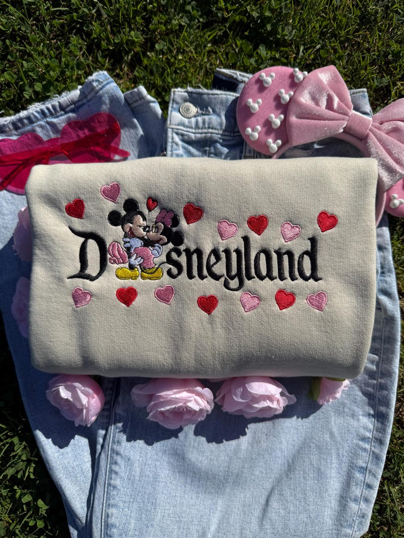 May include: A beige Disneyland sweatshirt with embroidered Mickey and Minnie Mouse, surrounded by red and pink hearts. The sweatshirt is laid on top of blue jeans, with pink roses and a pink headband with Mickey Mouse shapes.