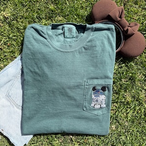 May include: A green t-shirt with a pocket featuring an embroidered image of R2-D2 from Star Wars. The t-shirt has a tag that reads "Comfort Colors" and "XL - TO 56".